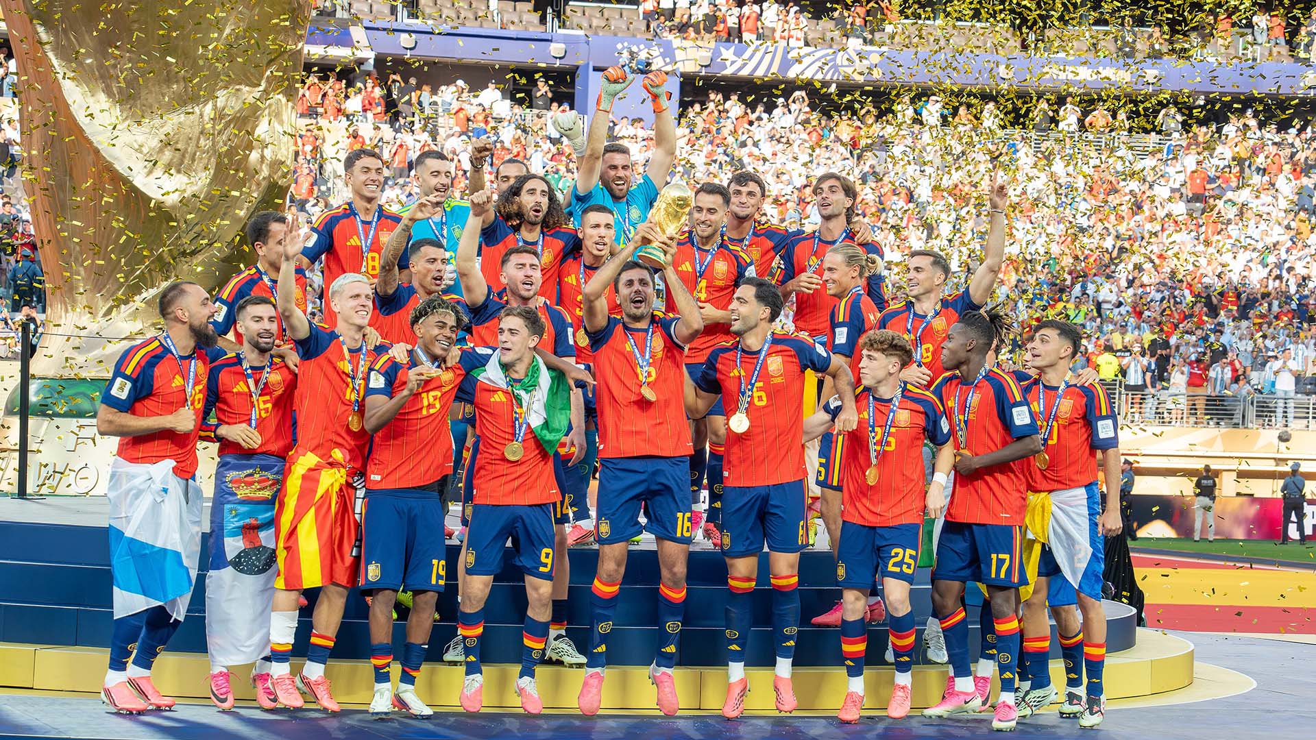 Jugadores de la sección española de fútbol, vestidos con el uniforme rojo y azul, celebran la victoria de la Copa Mundial de la FIFA 2026 sobre el podio del MetLife Stadium repleto de público, mientras cae confeti dorado alrededor. En el centro, el capitán de la sección Rodrigo Hernández Cascante (“Rodri”) levanta el trofeo dorado.