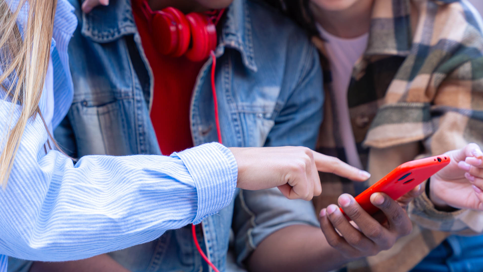 Tres adolescentes reunidos alrededor de un teléfono móvil rojo. Dos señalan la pantalla mientras uno de ellos (en el centro con camisa vaquera, camiseta y auriculares rojos al cuello) sostiene el dispositivo. La foto ilustra el acceso y la interacción con contenidos digitales y redes sociales de los adolescentes.