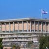 El edificio del Parlamento de Israel, conocido como la Knéset, situado en la cima de una colina en Jerusalén Oeste, coronado por una gran bandera de Israel ondeando en un mástil. El edificio está rodeado un aparcamiento de coches con señalizaciones de tráfico con una verja alta. Fuera de la verja, árboles y arbustos de la vía principal con banderas de Israel más pequeñas en los postes.