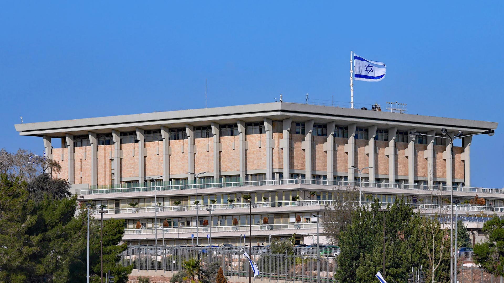 El edificio del Parlamento de Israel, conocido como la Knéset, situado en la cima de una colina en Jerusalén Oeste, coronado por una gran bandera de Israel ondeando en un mástil. El edificio está rodeado un aparcamiento de coches con señalizaciones de tráfico con una verja alta. Fuera de la verja, árboles y arbustos de la vía principal con banderas de Israel más pequeñas en los postes.