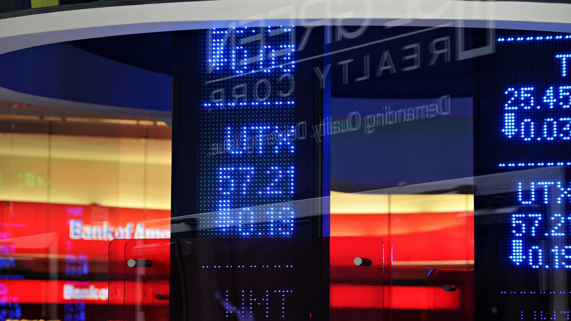Pantallas electrónicas con cotizaciones bursátiles y datos financieros iluminados en azul, reflejadas sobre superficies acristaladas. Al fondo, logotipo del Bank of America y los espacios interiores de la sucursal bancaria.