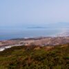 Vista panorámica desde una zona elevada de la frontera entre Ceuta y Fnideq (Castillejos). En primer plano aparecen colinas cubiertas de matorral y caminos de tierra; más abajo se observan carreteras, edificios e instalaciones próximas a la frontera. Al fondo, la ciudad se extiende junto a la costa, con el mar y varias montañas entre la bruma.