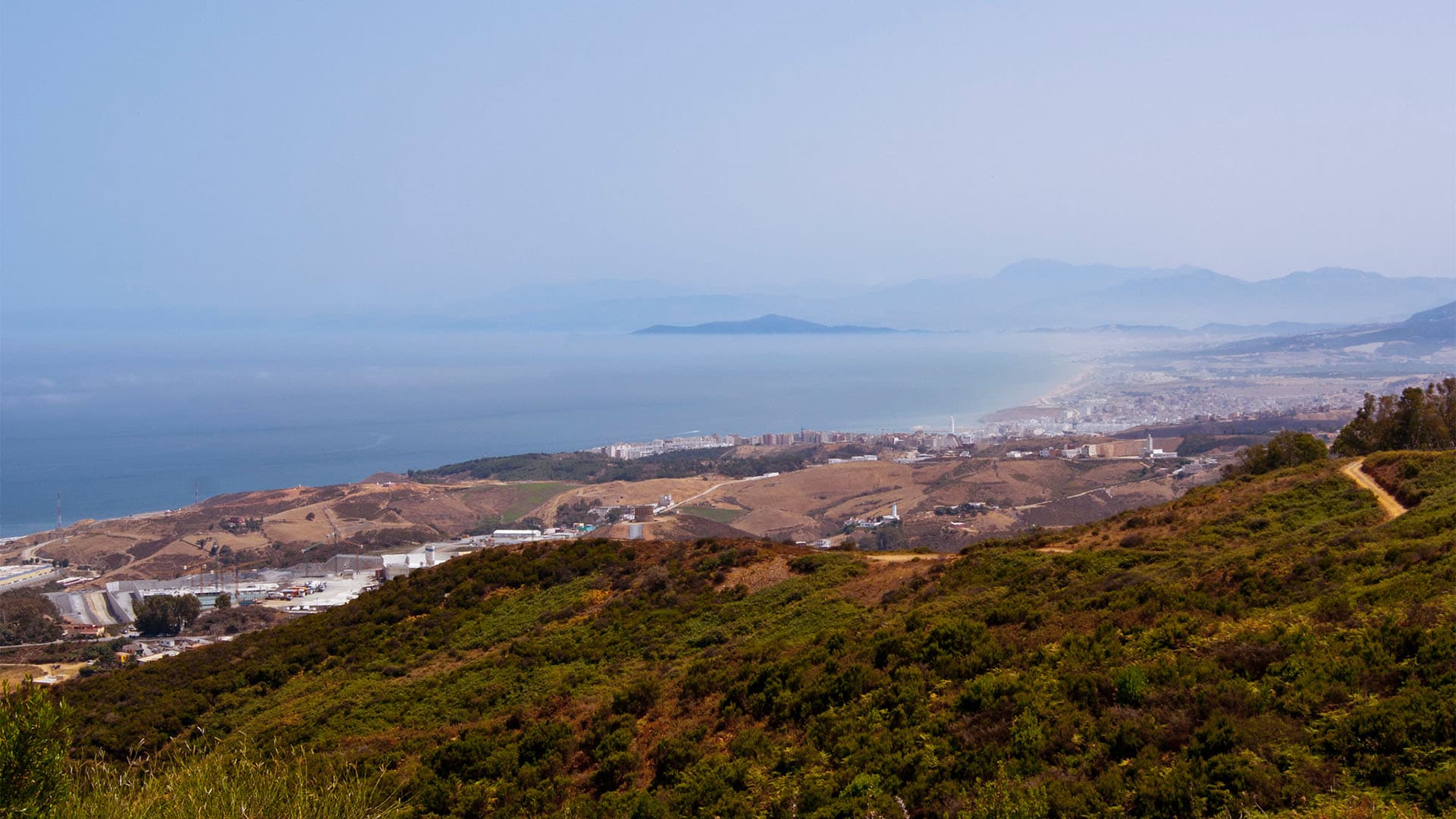 Vista panorámica desde una zona elevada de la frontera entre Ceuta y Fnideq (Castillejos). En primer plano aparecen colinas cubiertas de matorral y caminos de tierra; más abajo se observan carreteras, edificios e instalaciones próximas a la frontera. Al fondo, la ciudad se extiende junto a la costa, con el mar y varias montañas entre la bruma.
