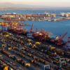 Vista aérea de los patios de contenedores y las terminales del puerto de Long Beach, en California, con filas de contenedores, grúas portuarias y varios buques en la bahía al atardecer.