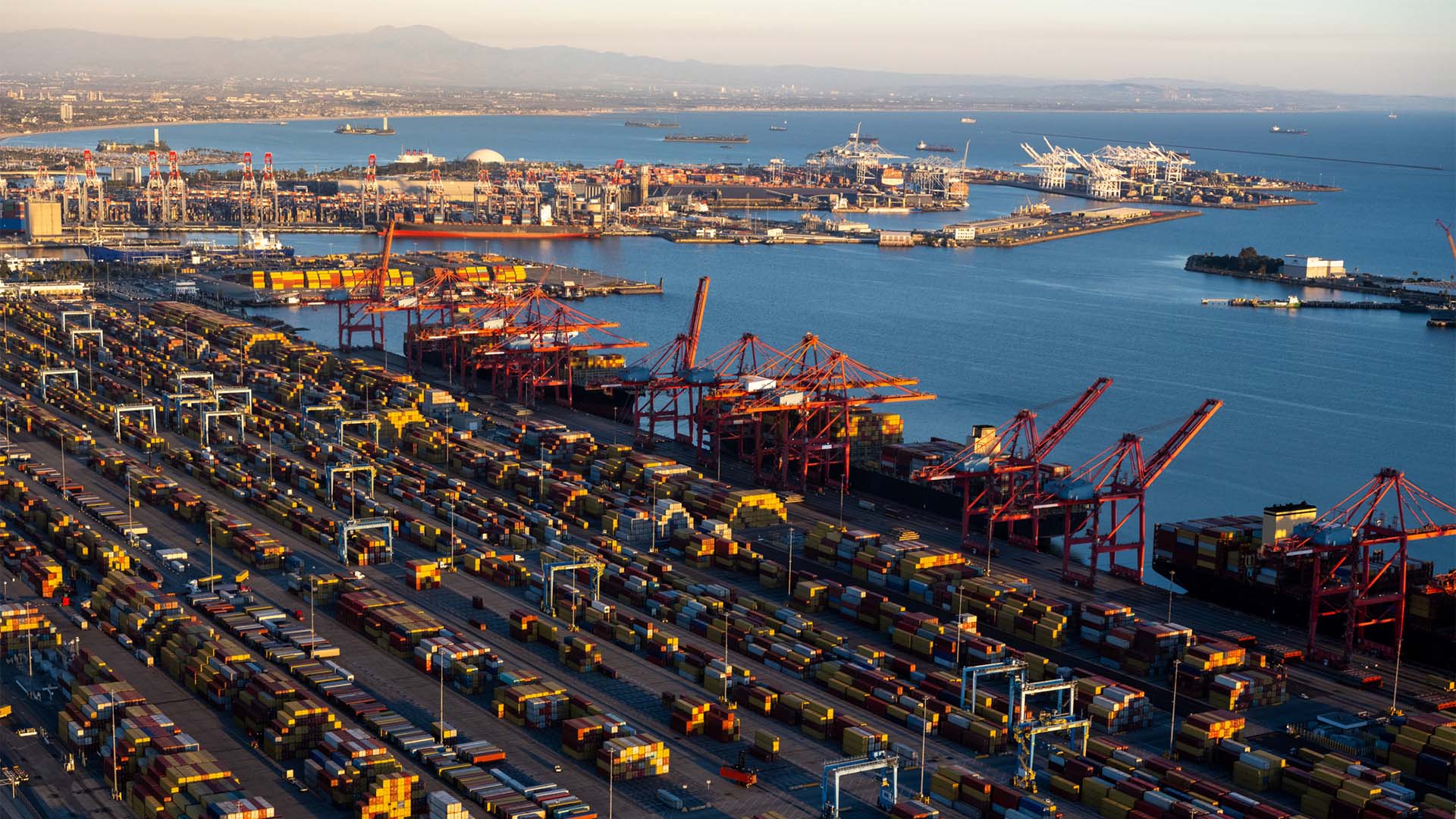 Vista aérea de los patios de contenedores y las terminales del puerto de Long Beach, en California, con filas de contenedores, grúas portuarias y varios buques en la bahía al atardecer.