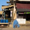 Mujer cubierta con un burka azul sentada frente a una tienda en Shor Bazaar, en el centro de Kabul, mientras varias personas caminan por la calle entre comercios y puestos.