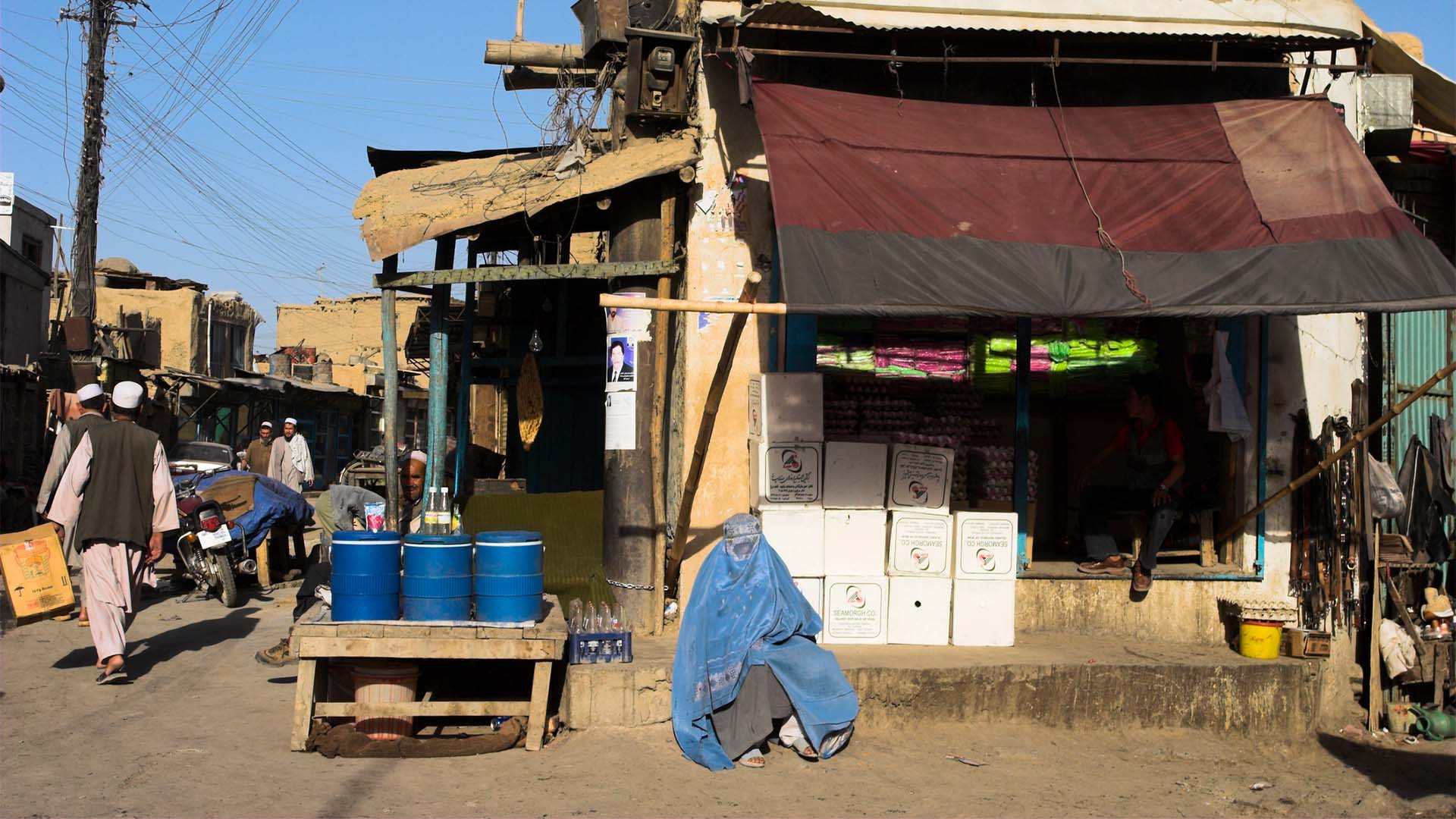 Mujer cubierta con un burka azul sentada frente a una tienda en Shor Bazaar, en el centro de Kabul, mientras varias personas caminan por la calle entre comercios y puestos.