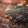 Crowds move through Jemaa el-Fna Square in Marrakech at sunset, surrounded by rows of food and market stalls with green canopies, as smoke rises from cooking areas and the evening light falls across the square.