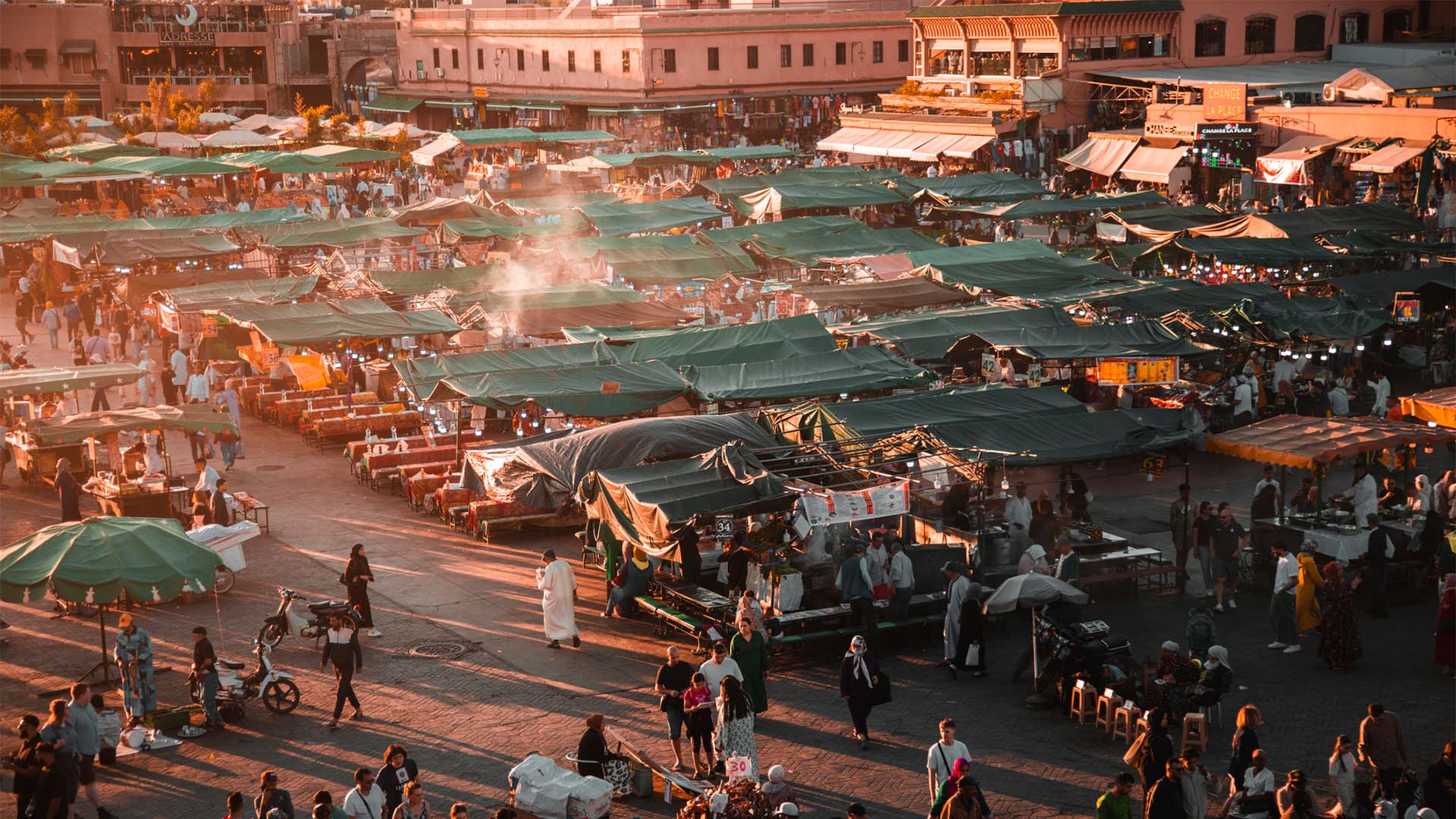 Crowds move through Jemaa el-Fna Square in Marrakech at sunset, surrounded by rows of food and market stalls with green canopies, as smoke rises from cooking areas and the evening light falls across the square.