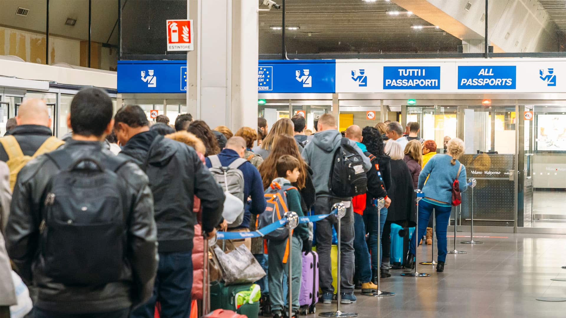 Numerosos viajeros con maletas y mochilas esperan en una larga fila ante los controles de pasaportes del aeropuerto de Milán-Malpensa, con carteles que indican el acceso para todos los pasaportes y viajeros extracomunitarios.