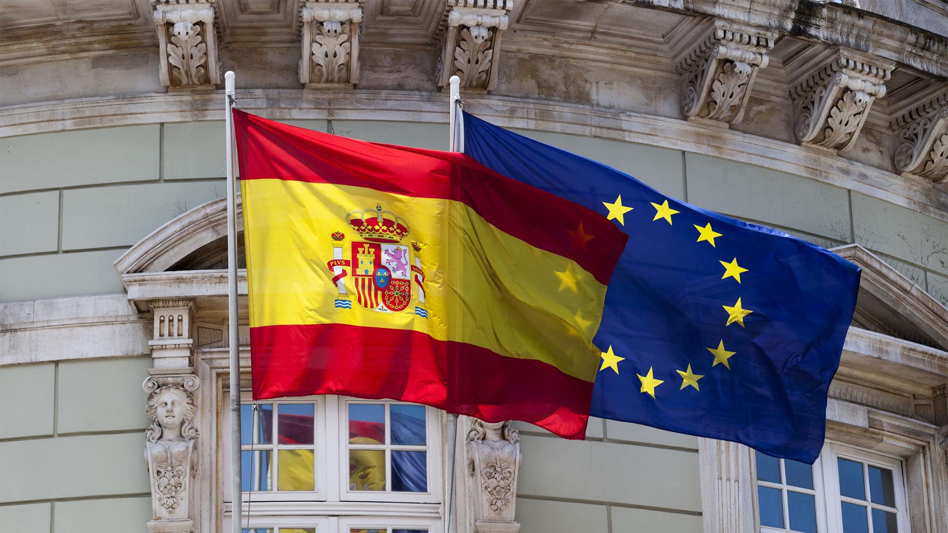 Banderas de España y de la Unión Europea ondeando juntas ante la fachada de un edificio institucional.