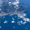 Vista aérea de de Ceuta y el Monte Hacho, con el núcleo urbano y el puerto junto a la costa, rodeados por las aguas azules del Mediterráneo y bajo nubes dispersas.