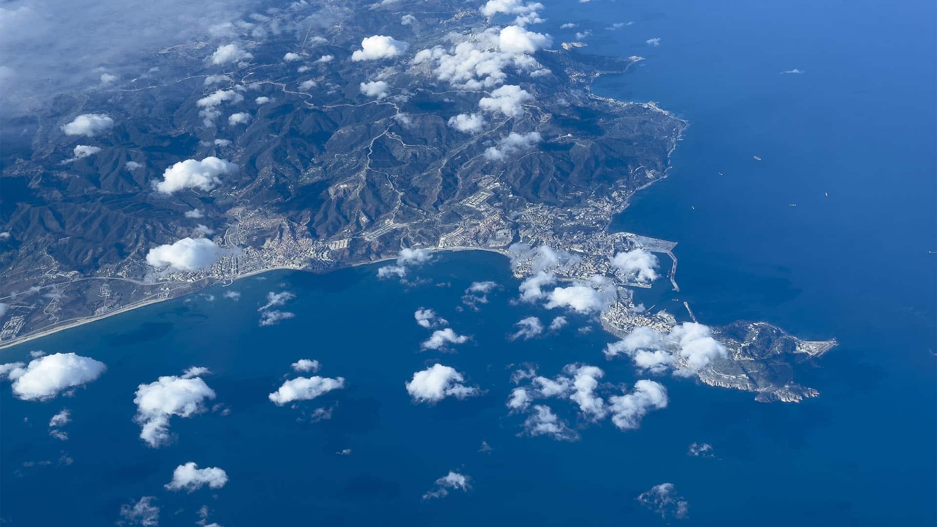 Vista aérea de de Ceuta y el Monte Hacho, con el núcleo urbano y el puerto junto a la costa, rodeados por las aguas azules del Mediterráneo y bajo nubes dispersas.
