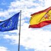 Las banderas de la Unión Europea y España ondean en un cielo claro y nublado. En la esquina inferior derecha, una palmera.