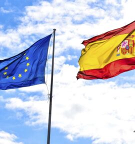 Las banderas de la Unión Europea y España ondean en un cielo claro y nublado. En la esquina inferior derecha, una palmera.