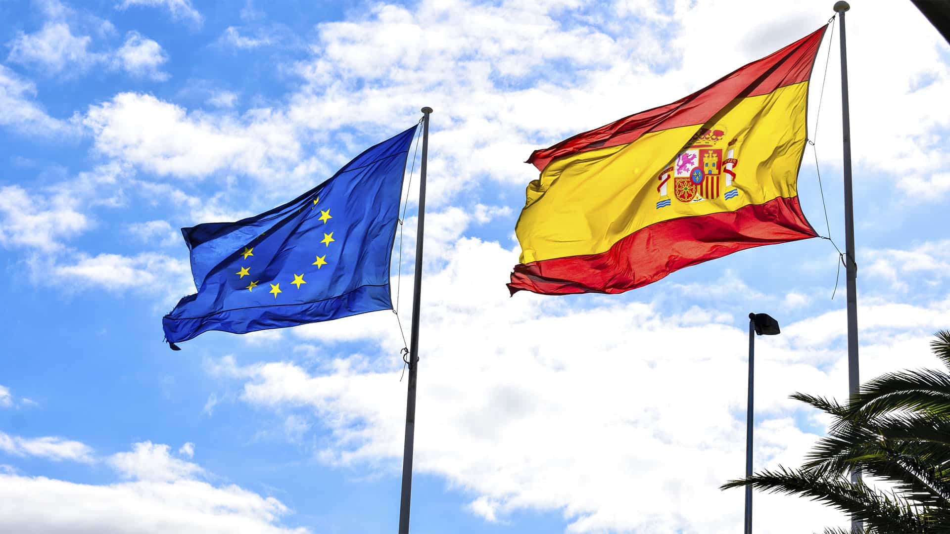 Las banderas de la Unión Europea y España ondean en un cielo claro y nublado. En la esquina inferior derecha, una palmera.