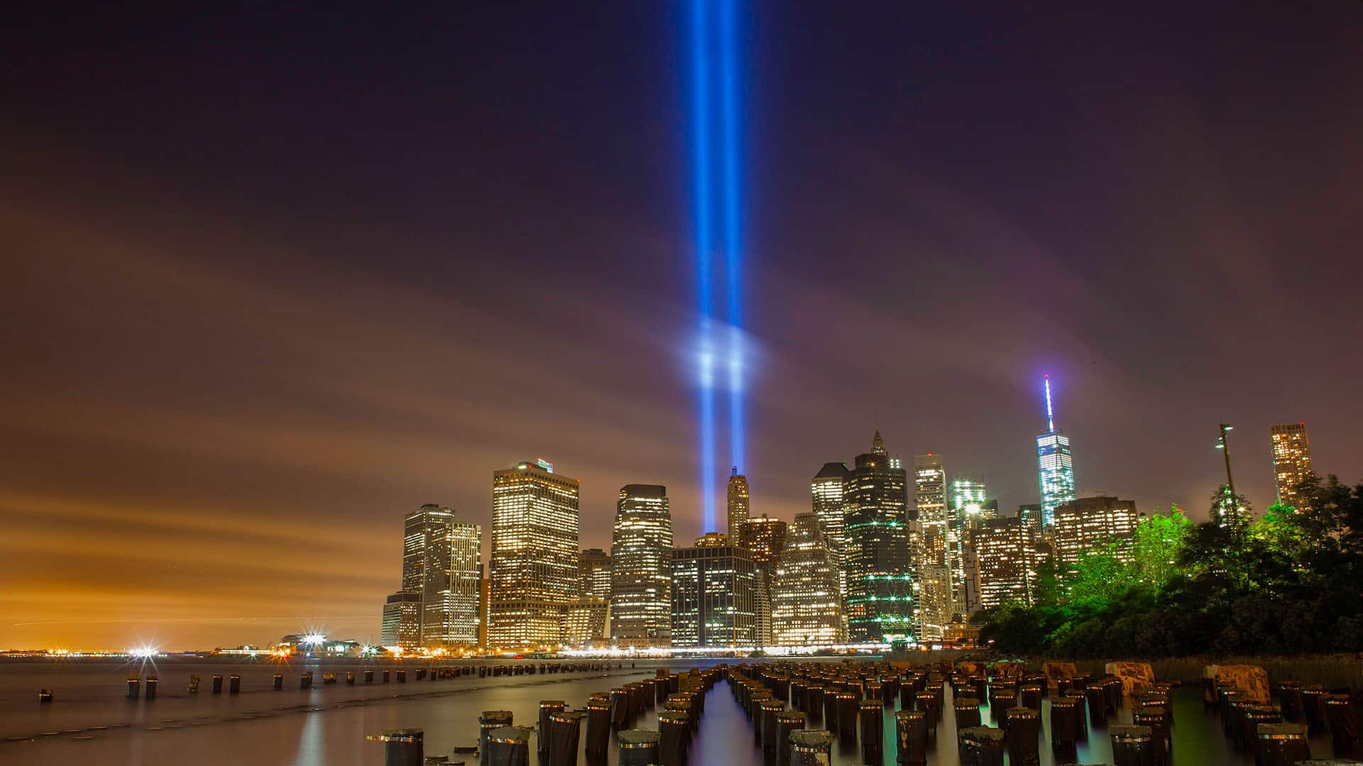 Horizonte urbano de Nueva York noche durante la instalación artística “Tribute in Light” en memoria de los atentados del 11-S, con dos haces de luz azul verticales proyectados al cielo desde los rascacielos junto al paseo marítimo.