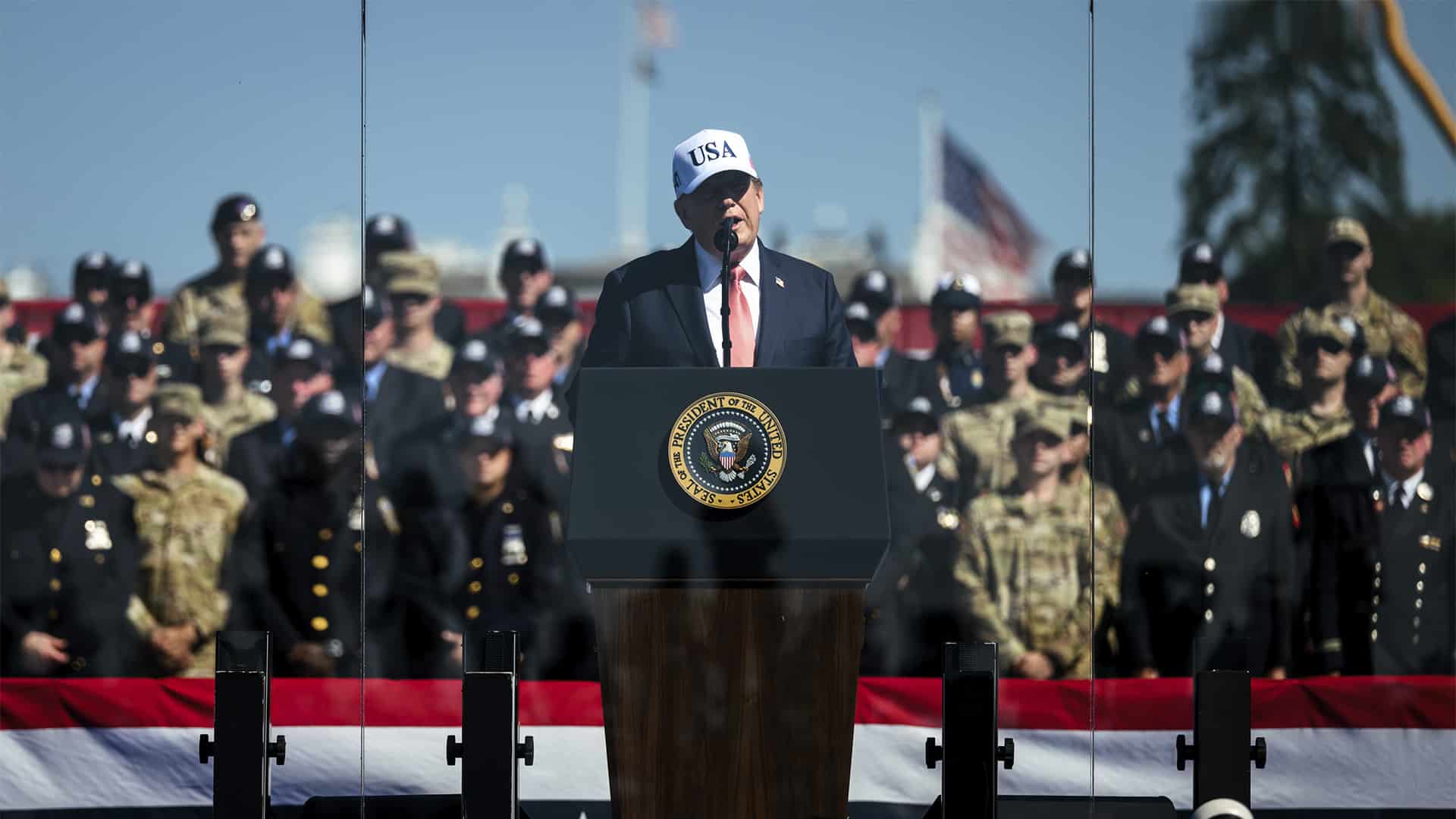 Donald Trump interviene ante un atril con el sello presidencial durante un acto conmemorativo del 25 aniversario del 11S al aire libre en Washington, rodeado de miembros de las fuerzas de seguridad y del Ejército, con banderas de Estados Unidos al fondo.