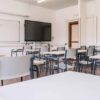 Bright, empty secondary school classroom in Madrid with desks arranged in pairs, grey chairs, white walls, ceiling lights, large whiteboards and a black digital screen at the front.