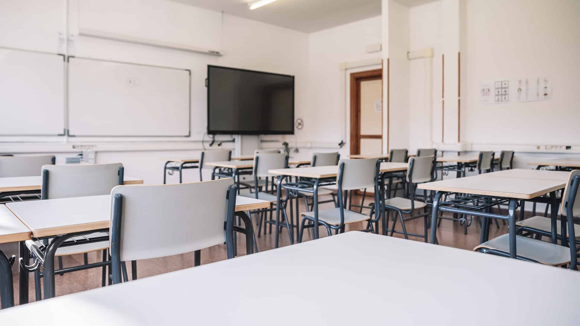Bright, empty secondary school classroom in Madrid with desks arranged in pairs, grey chairs, white walls, ceiling lights, large whiteboards and a black digital screen at the front.