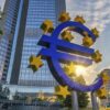 Escultura azul del símbolo del euro rodeada de estrellas amarillas frente a la sede del Banco Central Europeo en Fráncfort, con edificios de oficinas y el sol al fondo.