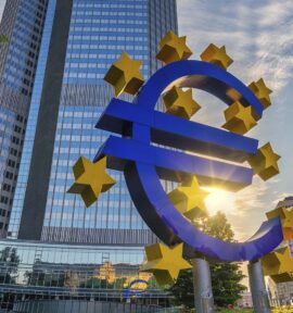 Escultura azul del símbolo del euro rodeada de estrellas amarillas frente a la sede del Banco Central Europeo en Fráncfort, con edificios de oficinas y el sol al fondo.