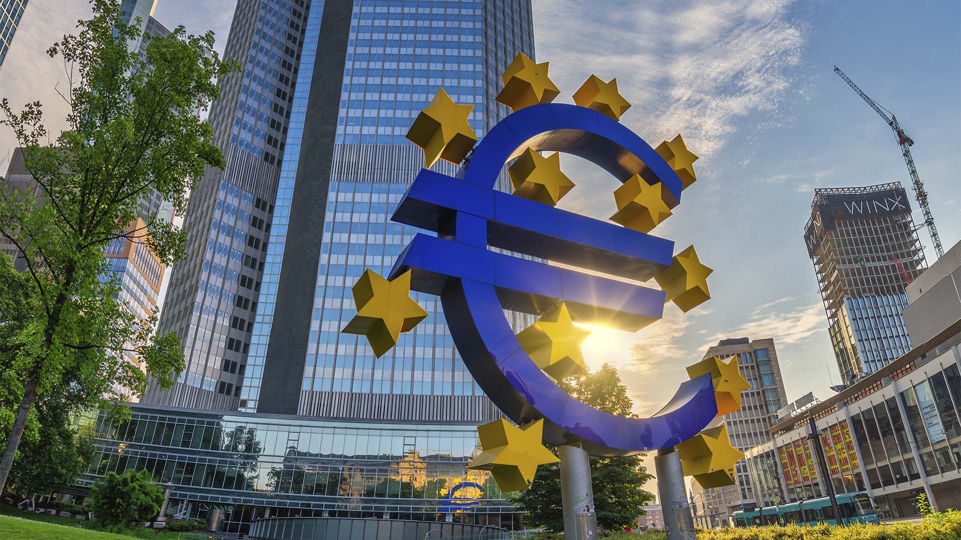 Escultura azul del símbolo del euro rodeada de estrellas amarillas frente a la sede del Banco Central Europeo en Fráncfort, con edificios de oficinas y el sol al fondo.