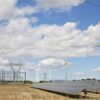 Solar panels and wind turbines beside an electrical substation and high-voltage power lines in Spain, under a partly cloudy sky.