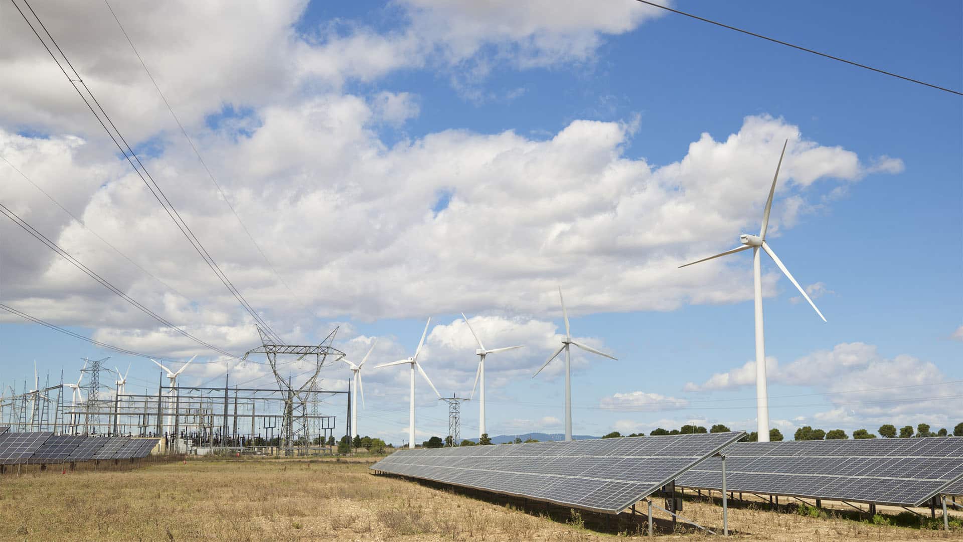 Solar panels and wind turbines beside an electrical substation and high-voltage power lines in Spain, under a partly cloudy sky.