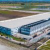 Centro de datos de Hutto, Texas (EE. UU.), visto desde el aire, con varios edificios principales de color blanco y gris, infraestructuras de refrigeración y una subestación eléctrica situada en la parte posterior del complejo. El campus está rodeado por un paisaje rural de campos abiertos.