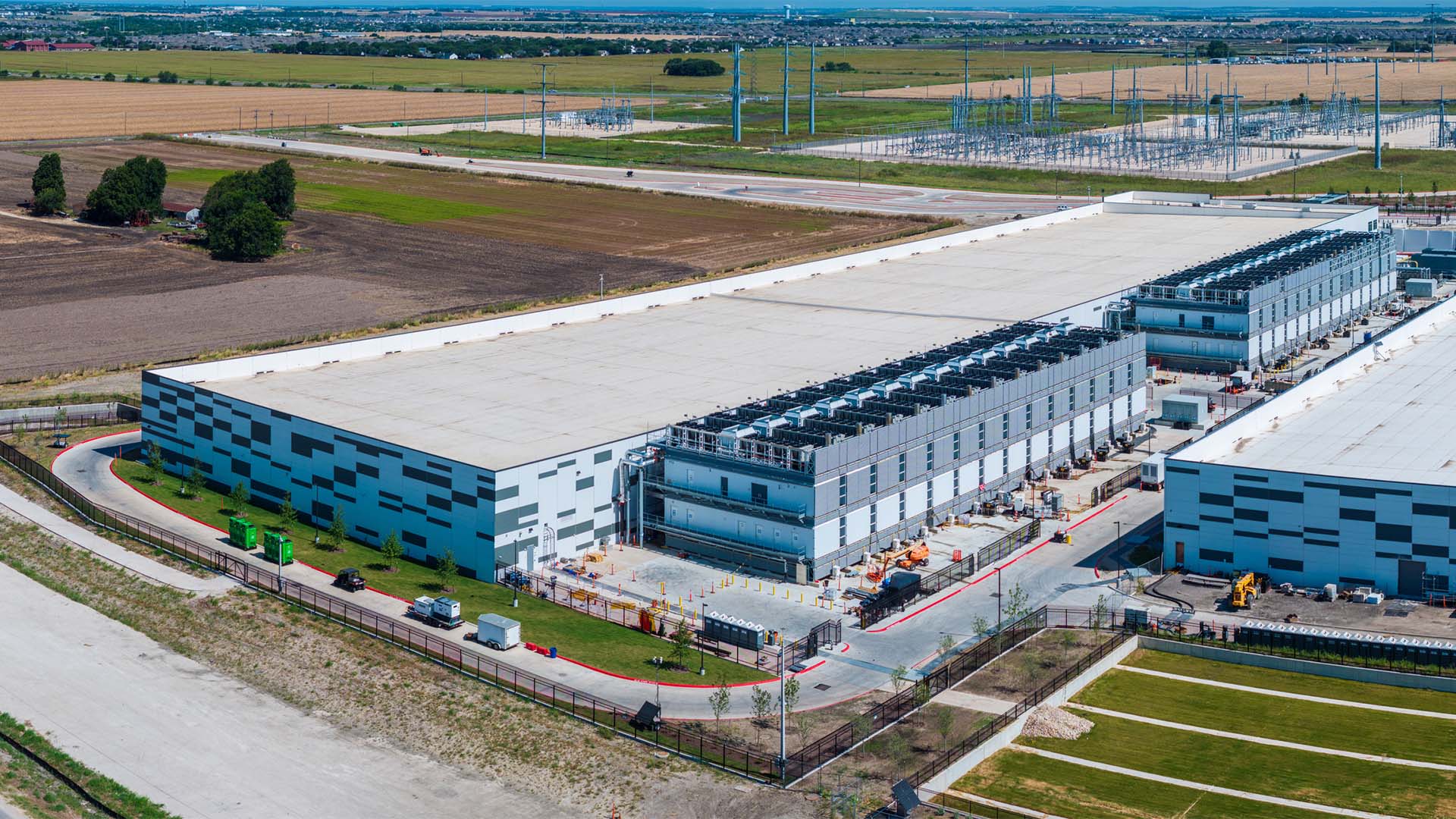 Centro de datos de Hutto, Texas (EE. UU.), visto desde el aire, con varios edificios principales de color blanco y gris, infraestructuras de refrigeración y una subestación eléctrica situada en la parte posterior del complejo. El campus está rodeado por un paisaje rural de campos abiertos.