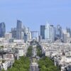 Vista panorámica de París con el distrito financiero de La Défense al fondo, formado por numerosos rascacielos y edificios de oficinas. En el centro destaca el arco de La Grande Arche, mientras en primer plano se extienden barrios residenciales, una amplia avenida arbolada y edificios urbanos bajo un cielo azul despejado.
