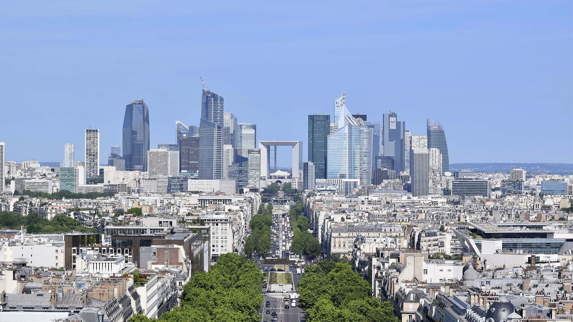 Vista panorámica de París con el distrito financiero de La Défense al fondo, formado por numerosos rascacielos y edificios de oficinas. En el centro destaca el arco de La Grande Arche, mientras en primer plano se extienden barrios residenciales, una amplia avenida arbolada y edificios urbanos bajo un cielo azul despejado.