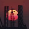 Silueta de una plataforma petrolífera y varias grúas frente a un gran sol rojo al atardecer en el estrecho de Ormuz.