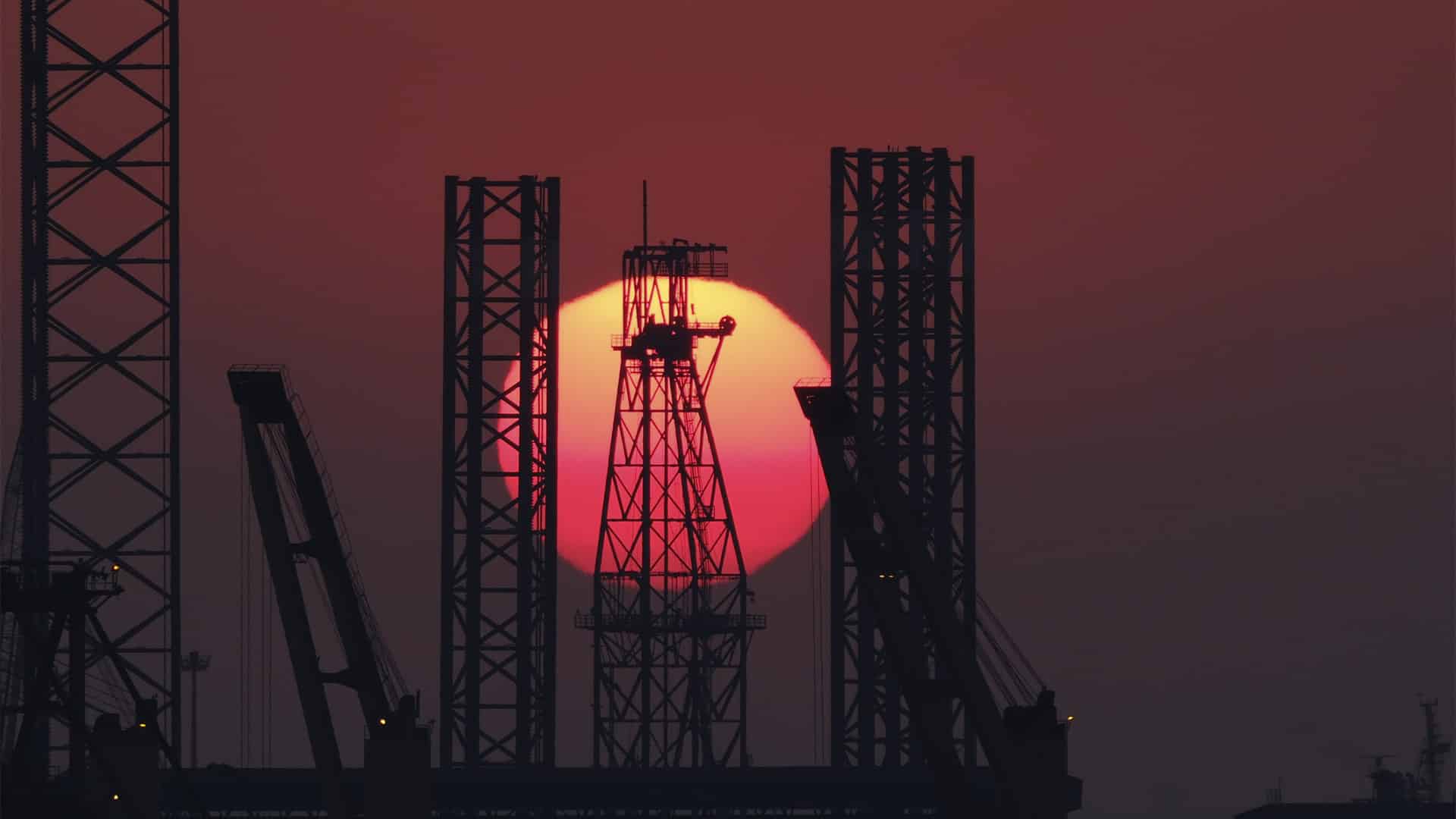 Silueta de una plataforma petrolífera y varias grúas frente a un gran sol rojo al atardecer en el estrecho de Ormuz.