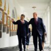 Xi Jinping y Donald Trump caminan juntos por una galería porticada de la Casa Blanca. Ambos señalan y observan una pared decorada con retratos enmarcados y placas conmemorativas, mientras varias personas de sus delegaciones caminan detrás de ellos.