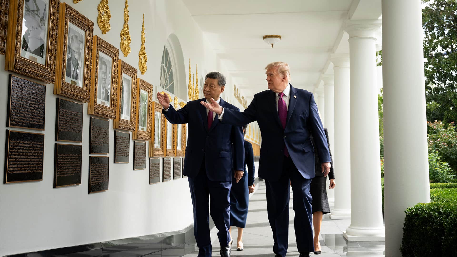 Xi Jinping y Donald Trump caminan juntos por una galería porticada de la Casa Blanca. Ambos señalan y observan una pared decorada con retratos enmarcados y placas conmemorativas, mientras varias personas de sus delegaciones caminan detrás de ellos.