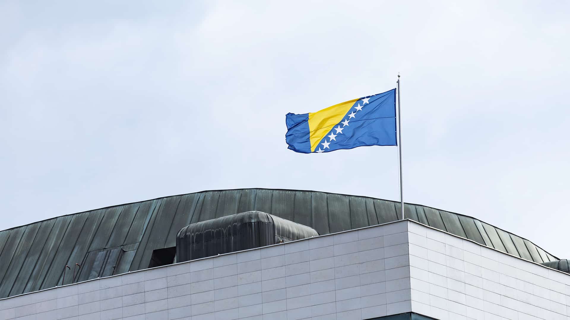 Bandera de Bosnia y Herzegovina ondeando sobre el edificio del Parlamento en Sarajevo, bajo un cielo nublado.