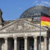 Vista frontal del edificio del Reichstag en Berlín, con su fachada de piedra y la gran cúpula de cristal al fondo. En primer plano ondea una bandera de Alemania de franjas negra, roja y amarilla, y otra bandera alemana aparece a la izquierda del edificio.