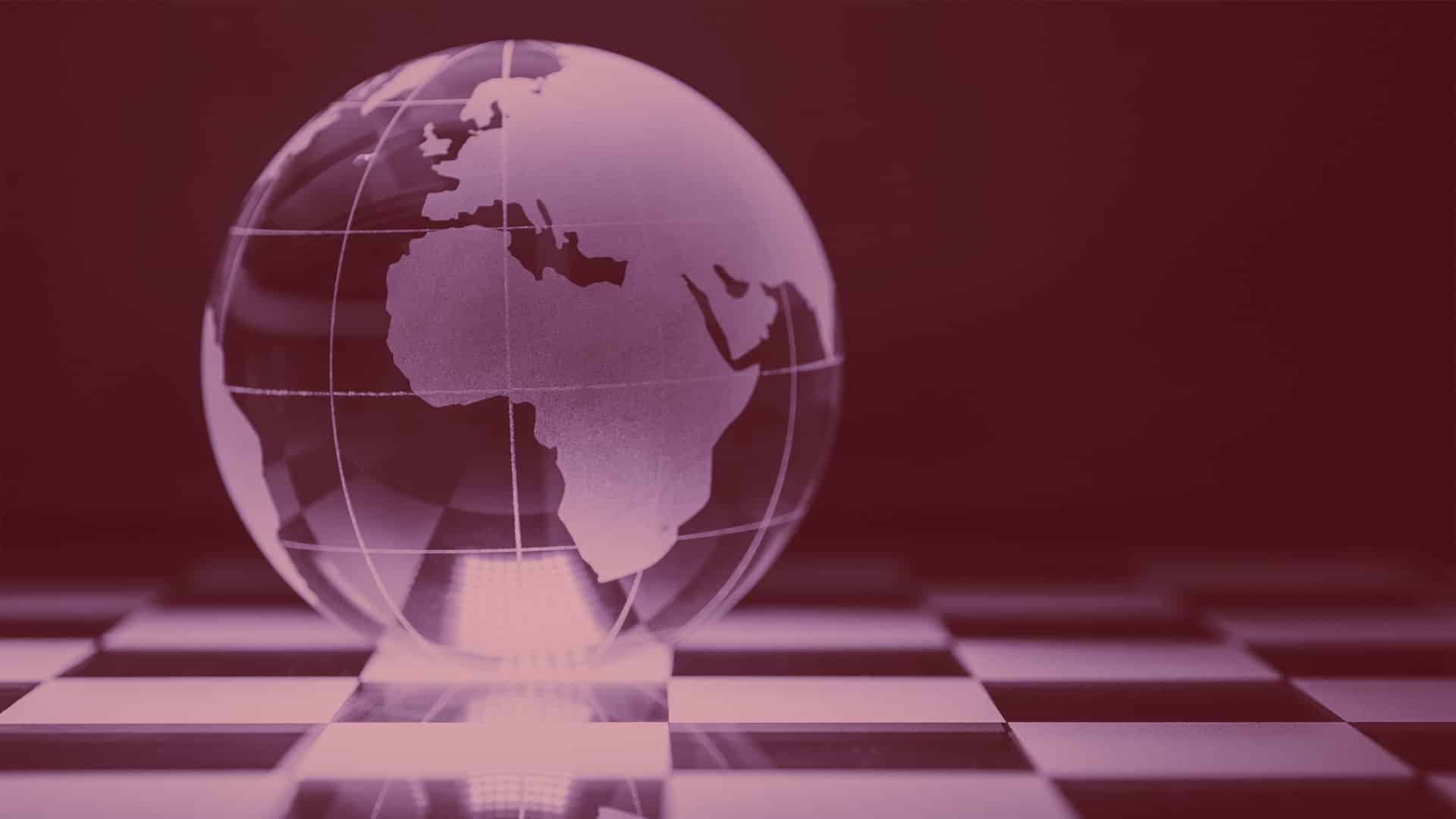 Globo terráqueo de cristal con Europa, África y parte de Asia grabadas, colocado sobre un tablero de ajedrez en tonos rojizos.
