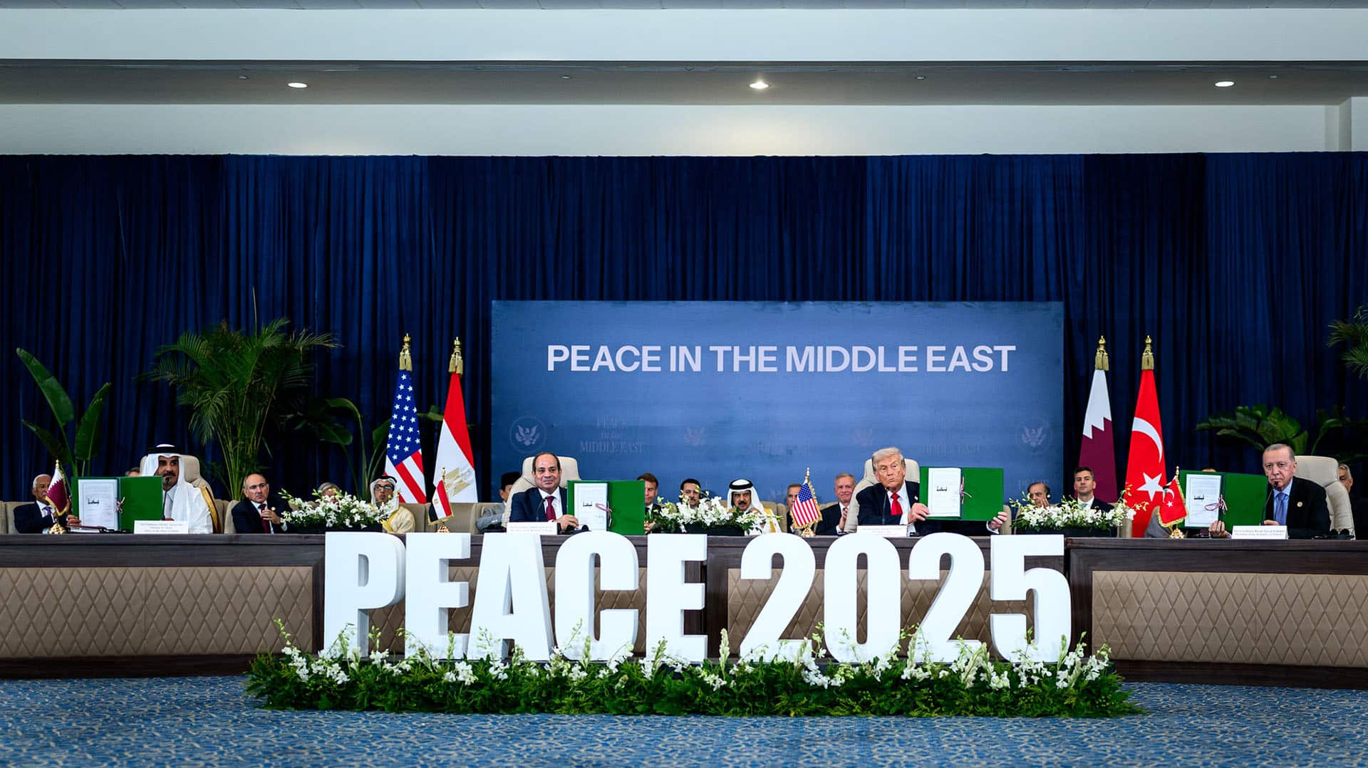 Ceremonia de firma del acuerdo de alto el fuego en la Cumbre en Sharm el-Sheikh (Egipto). En primer plano las palabras “Peace 2025”, en la mesa sentados sosteniendo las carpetas del acuerdo destacan Abdelfatah al Sisi (presidente de Egipto) y Donald Trump (presidente de EEUU). Al fondo las banderas de EEUU, Egipto, Qatar y Turquía, el resto de lideres mundiales, y una pantalla con las palabras “Peace in the Middle East”.