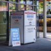 Buzón metálico gris y blanco para depositar papeletas de voto por correo del Distrito de Columbia, situado frente a un edificio acristalado en Washington D. C. Incluye inscripciones en inglés y español, entre las que destaca "Board of Elections. Official Ballot Drop Box". A su lado izquierdo, un cartel azul con letras blancas indica "Official Mail-In Ballot Drop Box" e incorpora los logotipos de Board of Elections y Vote Safe DC.