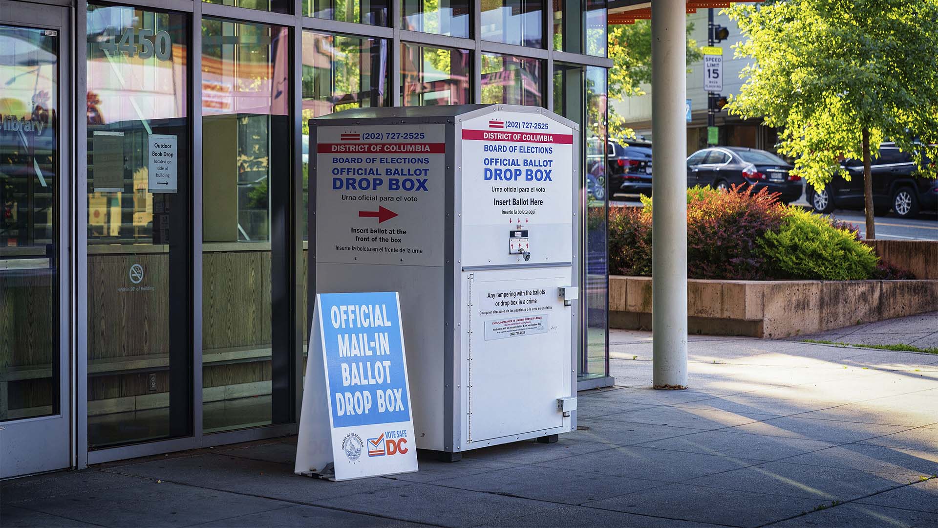Buzón metálico gris y blanco para depositar papeletas de voto por correo del Distrito de Columbia, situado frente a un edificio acristalado en Washington D. C. Incluye inscripciones en inglés y español, entre las que destaca "Board of Elections. Official Ballot Drop Box". A su lado izquierdo, un cartel azul con letras blancas indica "Official Mail-In Ballot Drop Box" e incorpora los logotipos de Board of Elections y Vote Safe DC.