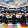 Vista general de los líderes europeos reunidos alrededor de una gran mesa circular en la sala S3 del Edificio Europa, en Bruselas, durante una sesión del Consejo Europeo. La sala presenta un suelo y un techo decorados con motivos geométricos de colores. Entre los asistentes se encuentran representantes políticos y responsables de los países miembros y representantes de los órganos de la UE.