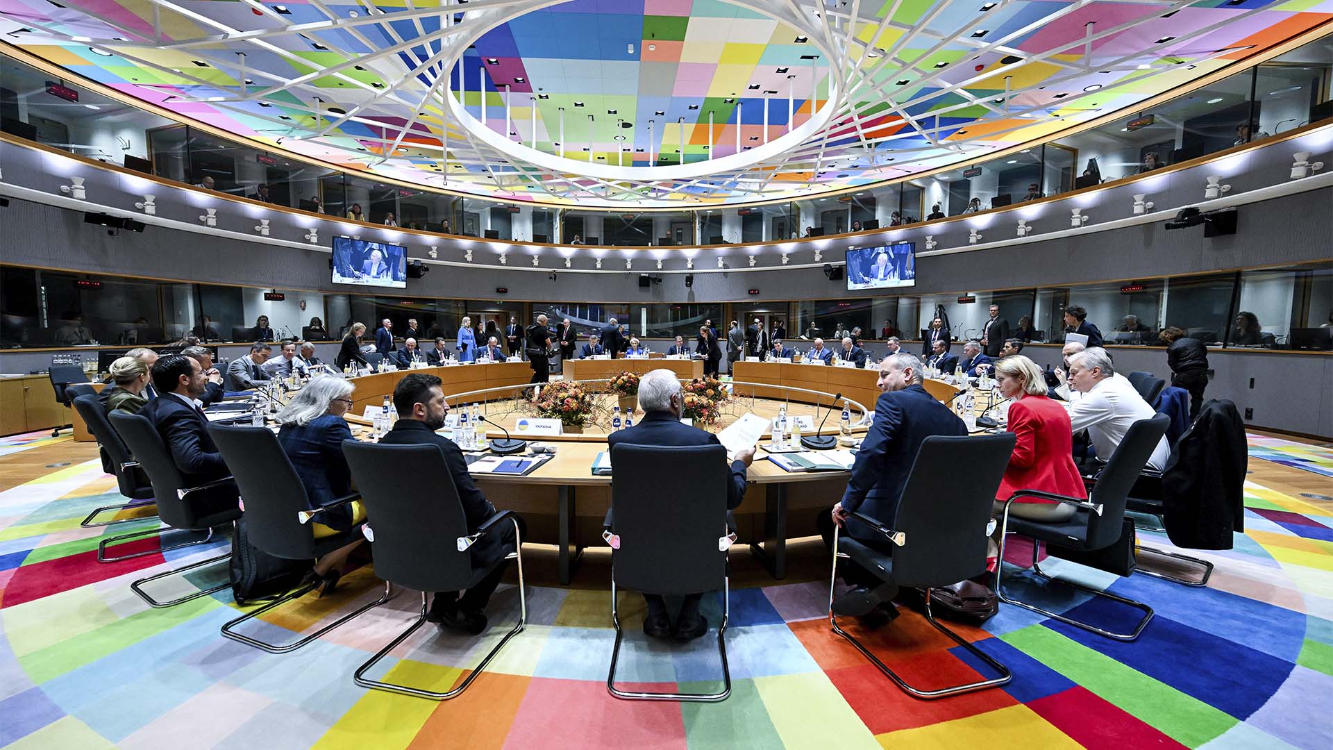 Vista general de los líderes europeos reunidos alrededor de una gran mesa circular en la sala S3 del Edificio Europa, en Bruselas, durante una sesión del Consejo Europeo. La sala presenta un suelo y un techo decorados con motivos geométricos de colores. Entre los asistentes se encuentran representantes políticos y responsables de los países miembros y representantes de los órganos de la UE.