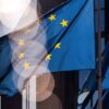 Varias banderas azules de la Unión Europea, con estrellas amarillas, ondean frente a la fachada acristalada de un edificio. La luz del sol ilumina parcialmente las banderas y produce reflejos circulares sobre la imagen.