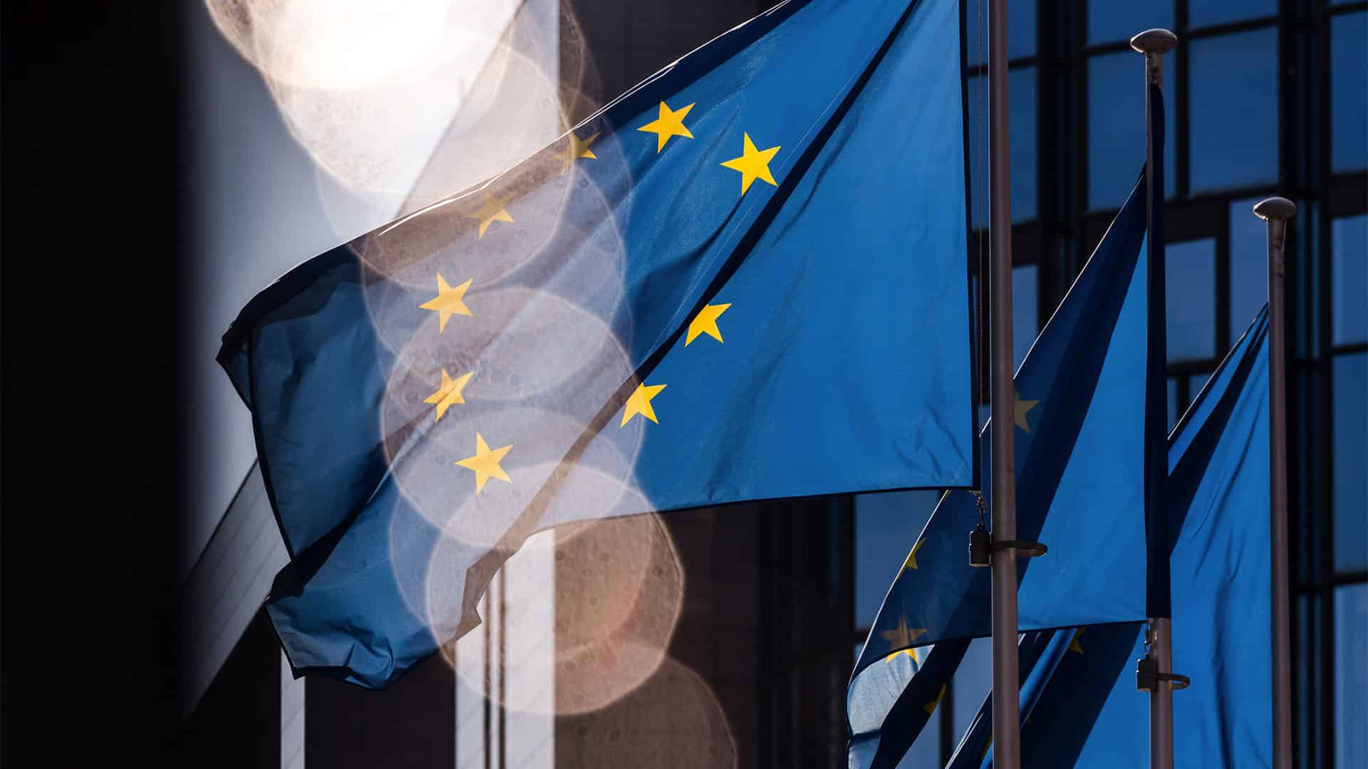 Varias banderas azules de la Unión Europea, con estrellas amarillas, ondean frente a la fachada acristalada de un edificio. La luz del sol ilumina parcialmente las banderas y produce reflejos circulares sobre la imagen.
