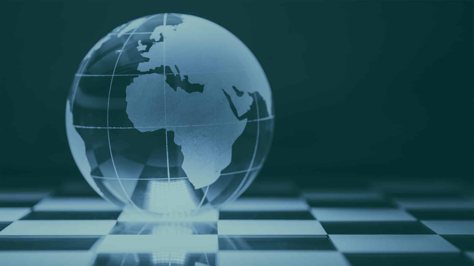 Globo terráqueo de cristal con Europa, África y parte de Asia grabadas, colocado sobre un tablero de ajedrez en tonos turquesas.