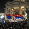 Multitud de simpatizantes de la Lista Estudiantil celebra la victoria electoral del 25 de octubre de 2026 frente a la Asamblea Nacional de Serbia, en Belgrado. Los asistentes ondean banderas serbias y se concentran de noche en las escalinatas del edificio, cuya fachada aparece iluminada. Una de las banderas aparece difuminada en primer plano, en el centro de la imagen.