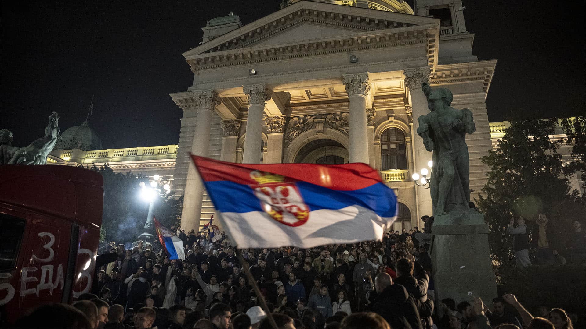Multitud de simpatizantes de la Lista Estudiantil celebra la victoria electoral del 25 de octubre de 2026 frente a la Asamblea Nacional de Serbia, en Belgrado. Los asistentes ondean banderas serbias y se concentran de noche en las escalinatas del edificio, cuya fachada aparece iluminada. Una de las banderas aparece difuminada en primer plano, en el centro de la imagen.